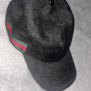 Svart Gucci keps med GG-mönster - Säljer en svart keps från Gucci med klassiskt GG-monogram över hela kepsen. Kepsen har en grön och röd rand på sidan, typisk för Gucci. Materialet är bomull och polyester, och modellen har böjd skärm och elastisk baksida för skön passform.