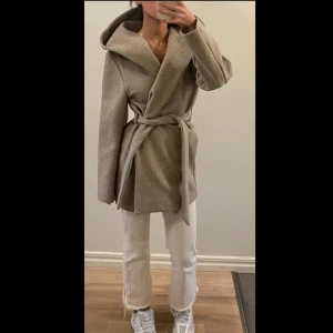 Beige omlott kappa från Only - Snygg beige kappa från Only med stor huva och omlottmodell. Jackan har ett bälte i midjan för att knyta och långa ärmar. Materialet är mjukt och känns som ullblandning, perfekt för höst och vinter. Enkel och stilren design som är lätt att matcha. Endast använd 2 gånger