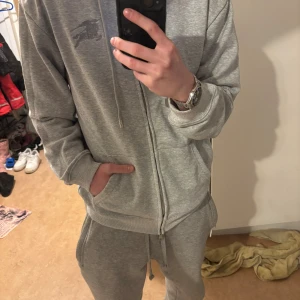 Grå hoodie från Burberry med dragkedja - Säljer en grå hoodie från Burberry i storlek M. Tröjan har dragkedja, snörning vid huvan och Burberrys klassiska broderade logga på bröstet. Insidan av huvan har det ikoniska rutiga Burberry-mönstret. Tillverkad i 100% bomull för skön komfort.