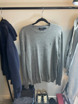 Grå stickad tröja Polo Ralph Lauren - Slim fit grå stickad tröja från Polo Ralph Lauren i mjuk merinoull. Klassisk rund hals och diskret broderad logga på bröstet. Långärmad modell med ribbade muddar och stilren look, perfekt för dig som gillar enkel och clean stil.