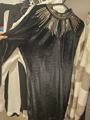 Svart långklänning med guldpaljetter - Svart långklänning i mjuk sammet med lång ärm och hög hals. Halsen är dekorerad med guldpaljetter i ett coolt strålande mönster. Klänningen har en elegant siluett och stängs med dragkedja bak. Perfekt för att sticka ut på festen.