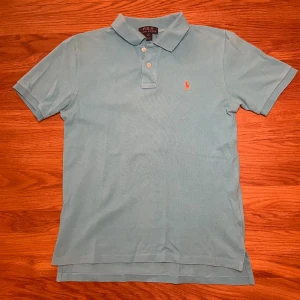 Ljusblå pikétröja från Polo Ralph Lauren - Snygg ljusblå pikétröja från Polo Ralph Lauren i klassisk modell med krage och tre knappar. Tröjan har korta ärmar och den ikoniska broderade loggan i rött på bröstet. Tillverkad i mjuk bomull som är skön mot huden och passar perfekt till vår och sommar.