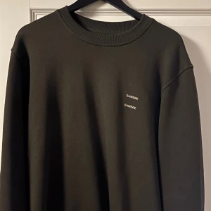 Mörkgrön sweatshirt från Samsøe Samsøe - En stilren mörkgrön sweatshirt från Samsøe Samsøe med rundad hals och diskret logga tryckt på bröstet. Tröjan har långa ärmar och ribbad kant vid halsen. Perfekt för dig som gillar minimalistisk design och vill ha en enkel, snygg look.