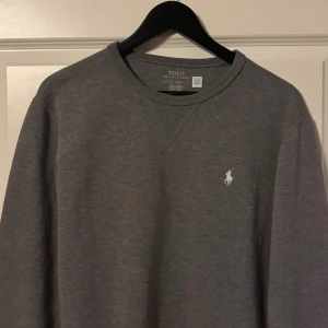 Grå sweatshirt från Polo Ralph Lauren - Snygg grå sweatshirt från Polo Ralph Lauren med klassisk rund hals och långärmad design. Tröjan har en liten vit broderad logga på bröstet och är tillverkad i mjuk bomullsmix som är skön att bära. Perfekt för chill dagar och enkel att matcha med jeans.