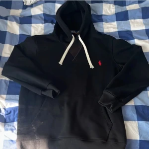 Svart hoodie från Polo Ralph Lauren - Svart hoodie från Polo Ralph Lauren med klassisk röd logobrodyr på bröstet. Tröjan har vit snörning i huvan, ribbade muddar och en stor magficka. Materialet är mjuk bomull och polyester, perfekt för chill dagar. Snygg och enkel design med en loose passform. Byten tas emot 