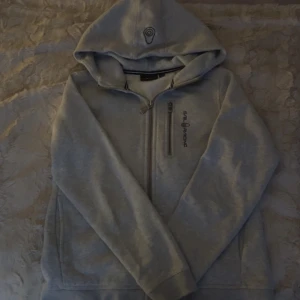 Grå hoodie från Sail Racing med dragkedja - Snygg ljusgrå hoodie från Sail Racing med huva och broderad logga på bröstet och huvan. Tröjan har hel dragkedja framtill, en extra bröstficka med dragkedja.