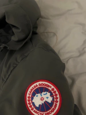 Grå canada goose jacka  - Säljer en grå jacka  från Canada Goose i storlek M då den inte passar mig längre. Jackan har huva, klassisk logga på ärmen och dragkedja med knappar framtill. Fylld med dun och har två sidofickor. Jackan är i perfekt skick 