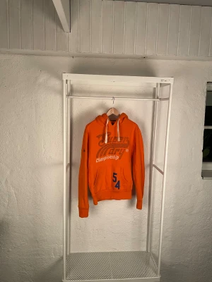 Orange hoodie från Superdry, S - Säljer en orange hoodie från Superdry i storlek S. Tröjan har tryck med text och siffror på framsidan, vit snörning i huvan och en klassisk känguruficka. Materialet är mjukt och känns som bomull, perfekt för chill dagar. Fleece på insidan för extra komfort.