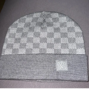 Grå rutig Louis Vuitton mössa - Snygg grå mössa från Louis Vuitton med klassiskt rutigt mönster och uppvikt kant. LV-logga i grått framtill ger en lyxig touch. Perfekt accessoar för dig som vill sticka ut med designerstil. Mjuk och stickad för skön komfort.