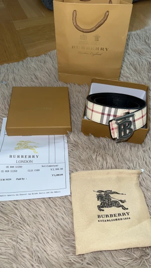 Burberry bälte - Snyggt Burberry-bälte med klassiskt rutigt mönster i beige, rött och svart. Bältet har ett stort metallspänne med Burberrys ikoniska B-logga. Utsidan är i mönstrat skinn och insidan är svart. Perfekt accessoar för att lyfta din outfit.