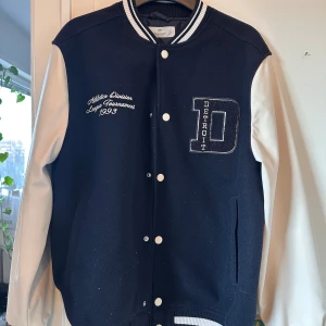 Svart och vit bomberjacka från H&M - Snygg svart och vit bomberjacka från H&M med broderad text och stort D för Detroit på bröstet. Jackan har vita ärmar i syntetmaterial, svart kropp i ullmix, ribbade muddar med ränder och knappar framtill. Perfekt för en sportig och trendig look.