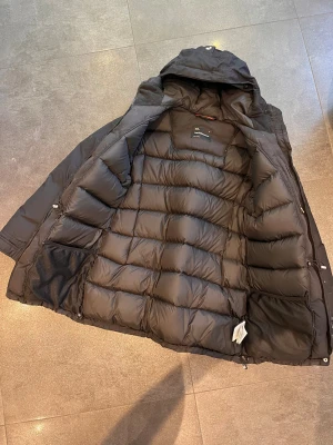 Svart dunjacka från Peak Performance - Säljer en svart dunjacka från Peak Performance med huva och flera praktiska fickor. Jackan har quiltad insida, dragkedja och knappstängning framtill. Perfekt för kalla dagar och har en clean, sportig look.