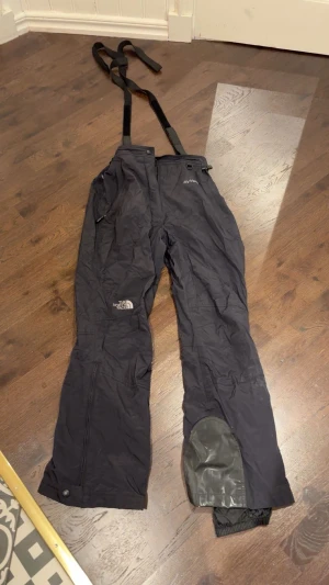 Svarta täckbyxor från The North Face - Svarta täckbyxor från The North Face med justerbara hängslen och förstärkning vid benslut. Byxorna har dragkedjor vid benen och är tillverkade i nylon med polyuretan och polyesterfoder. Perfekta för vintern och outdoor-aktiviteter.