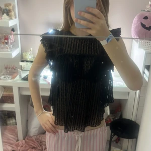 Svart spetsblus med volang från NELLY - Säljer en svart blus från NELLY med transparent spets och volangdetaljer vid axlarna. Blusen har korta ärmar, knytning i nacken och ett snyggt öppet ryggparti. Mönstret är randigt i spetsen och ger en cool vibe. Perfekt för dig som gillar statement-plagg!. Bara använd 1 gång❤️