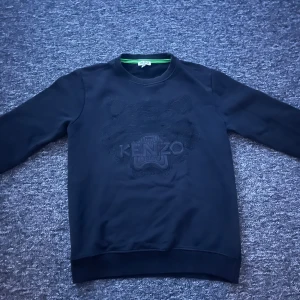Svart sweatshirt från Kenzo med tiger - Svart sweatshirt från Kenzo med det ikoniska tigermotivet broderat ton-i-ton på bröstet. Tröjan har rund hals, ribbade muddar och lång ärm. Perfekt för dig som gillar stilrena designer med en twist.