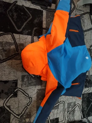 Färgstark vinterjacka Color Kids 140 - Cool vinterjacka från Color Kids i blått, mörkblått och orange. Jackan har huva, dragkedja och är tillverkad i vind- och vattentätt material med andasfunktion. Perfekt för kalla dagar och utomhusaktiviteter. Snygg och funktionell design för aktiva kids. Kommer också med en overall där nere de är båda för varann.