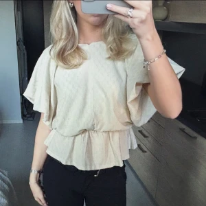 Beige blus med volang och kort ärm - Beige kortärmad blus/peplumtopp från NA-KD i storlek XS.  V-ringad i ryggen (ej djup). Använd en gång och inga deffekter så den är som ny🤍