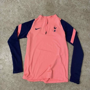 Tottenham långärmad fotbollströja Nike - Snygg långärmad fotbollströja från Nike med Tottenham Hotspur-logga. Tröjan är korallrosa med marinblå ärmar och detaljer, halv dragkedja framtill och sportig passform. Perfekt för träning eller match. Tillverkad i lätt och ventilerande material.