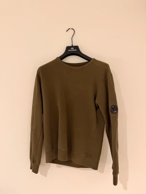 Olivgrön sweatshirt från C.P. Company - Snygg olivgrön sweatshirt från C.P. Company med rund hals och långärmad passform. Tröjan har deras ikoniska glaslins-detalj på vänster ärm och är gjord i mjuk bomull. Perfekt för dig som gillar stilrena och coola plagg. Köpt för ett år sedan för 3000 på nk. Tror jag har kvar kvittot. Passar folk mellan 160-170