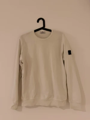 Beige sweatshirt från Stone Island - Säljer en stilren beige sweatshirt från Stone Island med klassisk rund hals och långärmad design. Tröjan har en svart patch med logga på vänster ärm och är tillverkad i mjuk bomull. Perfekt för dig som gillar clean och enkel streetwear. Passar folk mellan 165 till 173