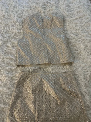 Champagnefärgad glittrig klänning H&M Divided - Champagnefärgad tvådelad klänning från H&M Divided med glittrigt paljettmönster i rutigt. Toppen är ärmlös och croppad, kjolen är kort och rak. Materialet är polyester och har en lyxig satinliknande känsla. Perfekt för dig som vill sticka ut med lite extra bling.