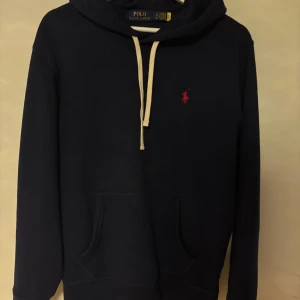 Mörkblå hoodie från Polo Ralph Lauren - Mörkblå hoodie från Polo Ralph Lauren med klassisk röd logobrodyr på bröstet. Tröjan har huva med beige snörning, känguruficka och långa ärmar. Perfekt för chill dagar och enkel att matcha med jeans eller joggers. Som ny: använd 2 gånger, nypris 2295kr. Mitt pris 1299kr
