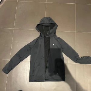 Svart vindjacka Peak Performance 160 - Svart vindjacka från Peak Performance med huva och dragkedja framtill. Jackan har två sidofickor med dragkedja och en liten logga på bröstet. Perfekt för dig som gillar stilren och sportig look. Materialet är syntet och jackan är långärmad.