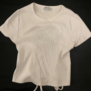 Vit ribbad topp med snörning - Säljer en vit ribbad topp från FB Sister (New horker) i storlek L. Men den sitter som en M/S. Toppen har korta ärmar och en rund halsringning. Baksidan har en snygg öppen rygg med snörningsdetalj som ger en cool och trendig vibe. Materialet är mjukt och stretchigt, perfekt för att skapa en chill look.