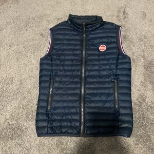 Mörkblå dunväst från Colmar - Snygg mörkblå dunväst från Colmar med quiltad design och två dragkedjeförsedda fickor. Västen har ett broderat Colmar-märke på bröstet och detaljer i rött, vitt och blått vid ärmöppningarna. Perfekt för lager-på-lager under kyliga dagar. Kan gå ner i pris vid snabb affär