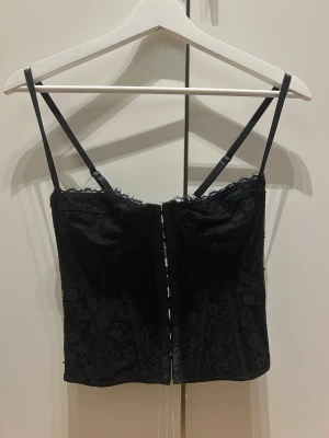 Svart spetskorsett med snörning - Säljer en svart korsett med blommig spets och smala axelband. Toppen har snygg snörning i ryggen och hakknäppning framtill. Materialet är mjukt och följsamt med transparent spetsdetaljer som ger en edgy vibe. Perfekt för dig som vill sticka ut.