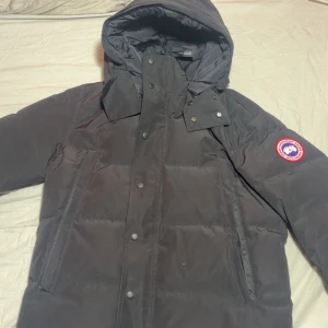 Svart dunjacka från Canada Goose - Säljer en svart dunjacka från Canada Goose med huva och klassisk logga på ärmen. Jackan har dragkedja och knappar framtill, två stora fickor och en stilren, puffig design som håller dig varm under kalla dagar.