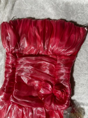 Röd axelbandslös volangklänning - Superfin röd axelbandslös kortklänning med glansigt tyg och volanger. Klänningen har en stor dekorativ rosett framtill och smockad rygg för skön passform. Perfekt för dig som vill sticka ut med en färgstark look och älskar detaljer. Helt ny 