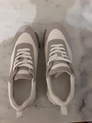 sneakers i vitt och grått - Säljer ett par sneakers i stilren design med vit och ljusgrå färg. Skorna har snörning, ribbad platt sula med grå detaljer och rund tå. Materialet är en mix av textil och mocka, med en cool H-logga på sidan och undersidan. Perfekta för dig som gillar exklusiva märken.Skick=10/10