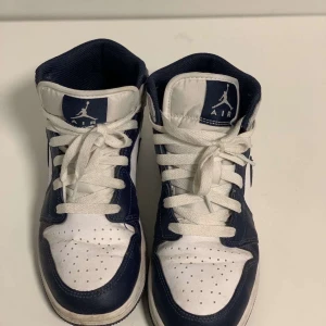 Nike Air Jordan 1 Mid Navy/Vit - Säljer ett par Nike Air Jordan 1 Mid i klassisk navy och vit färg. Skorna har snygg snörning, perforerad tå och ikoniska Jordan-loggor på sidan och plösen. Tillverkade i skinn och syntet med en platt sula och rund tå. Perfekta för dig som gillar streetstyle och sneakers med attityd.