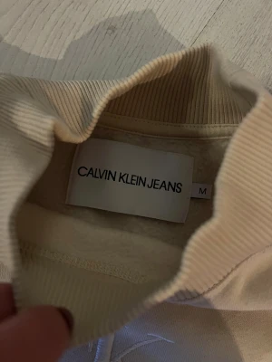 Beige sweatshirt från Calvin Klein Jeans - Snygg beige sweatshirt från Calvin Klein Jeans med broderad CK-logga framtill. Tröjan har ribbad hög krage, muddar vid ärmslut och nederkant samt långärmad passform. Perfekt för dig som gillar stilrena och klassiska plagg med en modern touch.