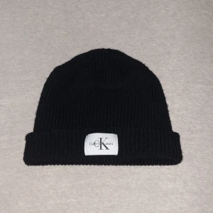 Svart ribbstickad mössa Calvin Klein - Svart ribbstickad mössa från Calvin Klein Jeans med uppvikt kant och vit logotyp-patch framtill. Tillverkad i 100% ull för extra värme och stilren look. Perfekt accessoar för kalla dagar och enkel att matcha med alla outfits.