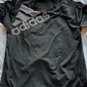 Svart Adidas t-shirt Freelift M - Svart t-shirt från Adidas med stort vitt logotryck på bröstet. Modellen är Freelift och tillverkad i lätt polyester med AEROREADY-teknologi som andas. Rund hals och korta ärmar, perfekt för träning. Snygg och sportig stil.