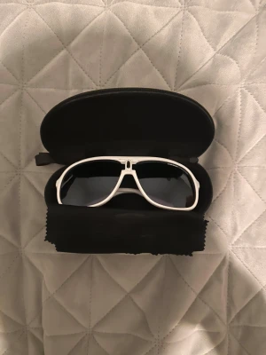 Vita Carrera pilotglasögon - Snygga vita pilotglasögon från Carrera med svarta tonade linser. Glasögonen har en klassisk aviatorform och kommer med ett svart hårt fodral och putsduk. Perfekt för dig som vill ha en cool och sportig look.