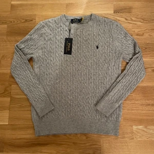 Grå kabelstickad tröja Polo Ralph Lauren - Snygg grå kabelstickad tröja från Polo Ralph Lauren med klassisk rund hals och broderad logga på bröstet. Tröjan har ribbade muddar och är långärmad, perfekt för dig som gillar stilrena och tidlösa plagg. Passar dig som vill ha en clean look.   OBS Storlek M men känns närmare S