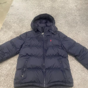 Mörkblå pufferjacka Polo Ralph Lauren  - Säljer en mörkblå pufferjacka från Polo Ralph Lauren med huva och dragkedja. Jackan har två fickor med dragkedja och klassisk röd Polo-logga på bröstet. Perfekt för kalla dagar och riktigt skön att bära. Använd en vinter och inte en ända skråma. Ska passa folk med XS i storlek, annars är det barn XL.