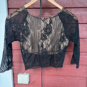 Svart spetsblus från Bik Bok XS - Säljer en svart blus från Bik Bok i storlek XS. Blusen är croppad med långärmade spetsärmar och har ett transparent mönster med både ränder och blommor. Den har en nude-färgad underdel som ger en snygg kontrast mot den svarta spetsen. Perfekt för dig som gillar detaljer!