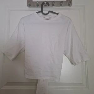 Vit omlottopp med knytband - Säljer en vit topp i bomull med trekvartsärm och snyggt knytband i ryggen. Modellen är croppad och har en enkel, stilren look som passar perfekt till jeans eller kjol. Omlott-detaljen bak ger en extra twist till outfiten.