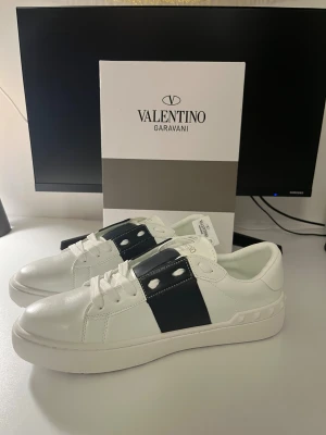 Valentino Garavani sneakers vit/mörkblå - Snygga Valentino Garavani sneakers i vitt skinn med bred svart rem över vristen och coola nitar runt hälen. Klassisk rund tå, platt sula och vita snören. Perfekta för dig som vill ha en stilren men ändå edgy look.