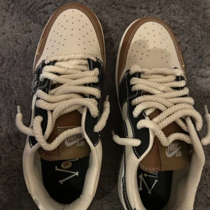 Nike Air Force 1 Low i beige och brunt - Low sneakers med beige och bruna paneler, svart Swoosh och detaljer i guld. Grova vita snören och perforerad tå för extra stil. Skorna har en platt sula och är tillverkade i skinn med kontrasterande sömmar och broderade detaljer på sidan. Kan ej identifiera äkthet. Knappt använda alls.