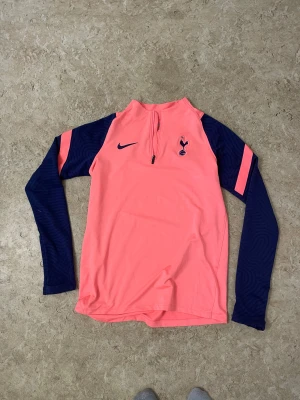 Nike Tottenham långärmad träningströja - Snygg långärmad träningströja från Nike x Tottenham Hotspur i en fräsch rosa färg med marinblå ärmar och detaljer. Tröjan har half zip framtill, klubbmärke på bröstet och är gjord i mjukt polyester-material som andas. Perfekt för dig som gillar fotboll och vill sticka ut på planen.