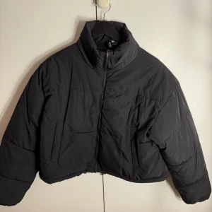 Svart croppad pufferjacka H&M Divided - Svart croppad pufferjacka från H&M Divided med hög krage och dragkedja framtill. Jackan har långa ärmar, två fickor och snörning i nederkant för att justera passformen. Perfekt för kalla dagar och har en clean, modern look.