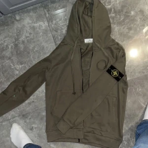 Olivgrön hoodie från Stone Island - Säljer en olivgrön hoodie från Stone Island med klassisk patch på vänster ärm. Tröjan har huva med snörning, dragkedja framtill och två fickor. Materialet är mjuk bomull och passformen är normal. Perfekt för dig som vill ha en Clean outfit.jag köpte den på plick för 1700 vet ej om den e äkta skriv vid frågor. Den e använd max 5 gånger 