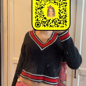 Svart croppad stickad tröja Divided - Säljer en svart croppad stickad tröja från Divided med ribbad struktur. Tröjan har v-ringad hals med röda och vita ränder samt matchande ränder vid midjan och ärmsluten. Långärmad och perfekt för dig som gillar sportig och trendig stil.