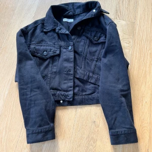 Svart croppad jeansjacka från Gina Tricot - Svart croppad jeansjacka från Gina Tricot med klassisk krage, bröstfickor med knappar och långa ärmar. Jackan har en snygg, rak passform och är tillverkad i kraftigt bomullstyg. Perfekt för dig som vill ha en stilren och enkel look. Köpt begagnad men passade tyvärr inte mig så säljer vidare den. Storlek XS 