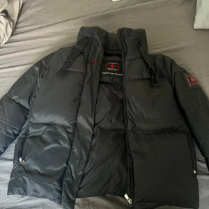 Svart pufferjacka från Canada Snow - Säljer denna snygga pufferjackan från Canada Snow med hög krage och broderad lönnlöv på nacken. Jackan har dragkedja, stora fickor och patch med logga på ärmen. Perfekt för kalla dagar och riktigt varm.